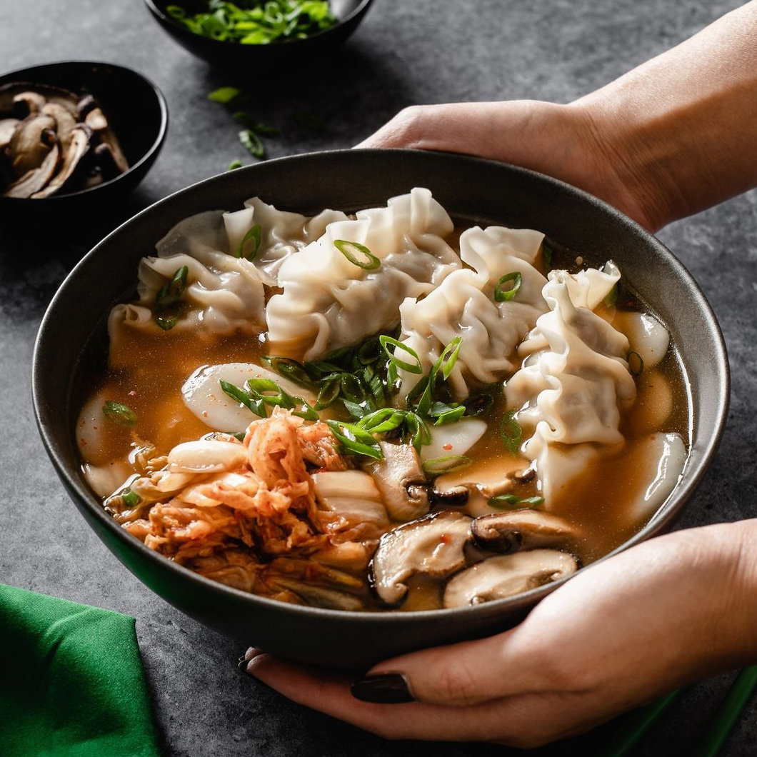 Eine Schale Tteok Mandu Guk, gefüllt mit Mandu (Teigtaschen), Tteok (Reiskuchen), Pilzen und Kimchi in einer klaren Brühe.