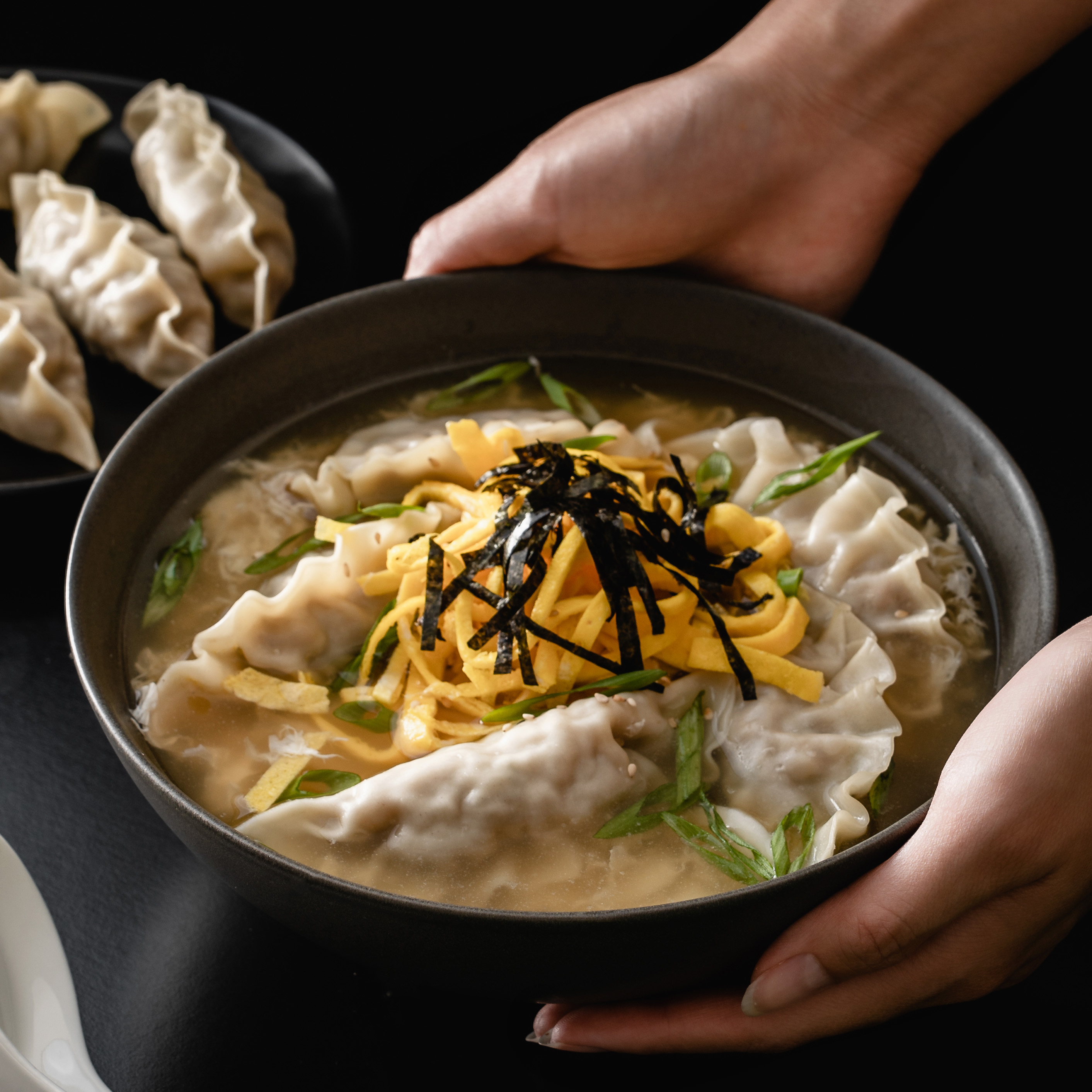 Eine Schale Mandu Guk mit bibigo Dumplings, garniert mit dünnen Eigelbstreifen, Algen, Sesamöl und Frühlingszwiebeln. Die herzhafte Brühe enthält Dumplings, Eistränge und eine reichhaltige Mischung aus Aromen.